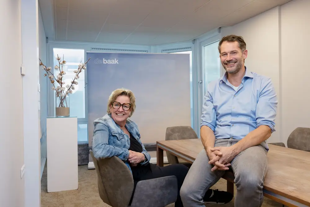 Een man en vrouw bij een vergadertafel de vrouw leunend op een stoel de man zitten op tafel
