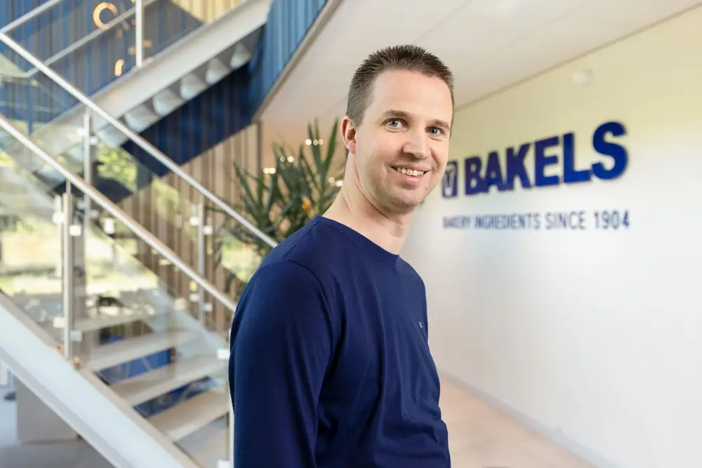 Nicky Gommers van Bakels staand voor het Logo van Bakels Senior