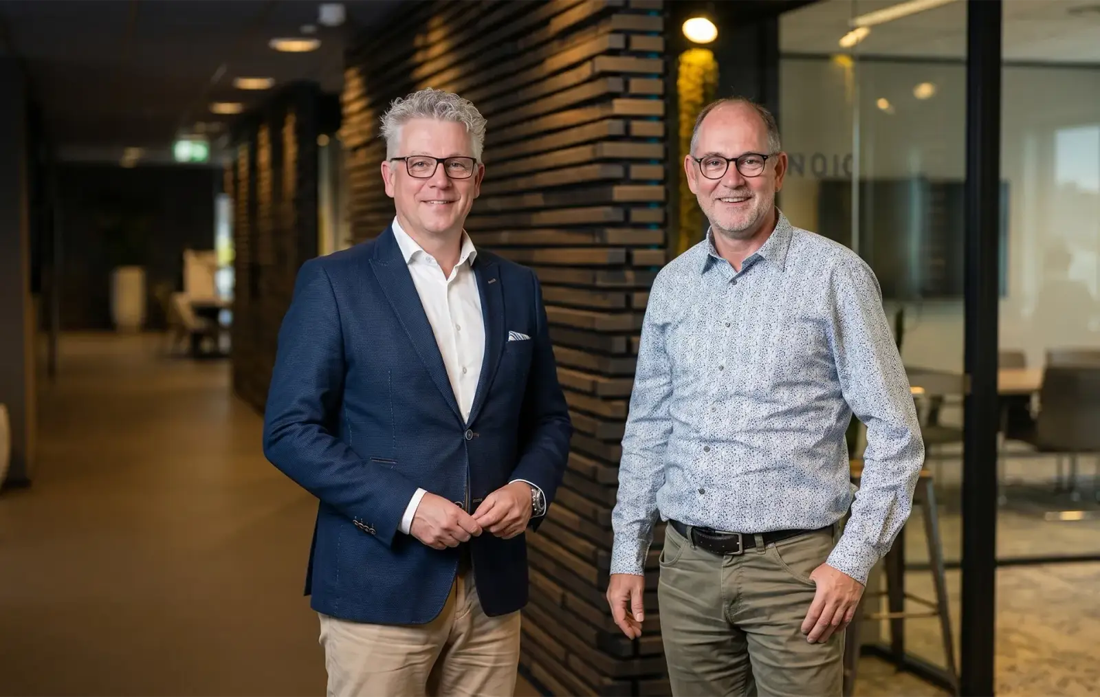 Collega's Michell en Harro van Axelio Academy staan lachend naast elkaar in de gang van een kantoorgebouw