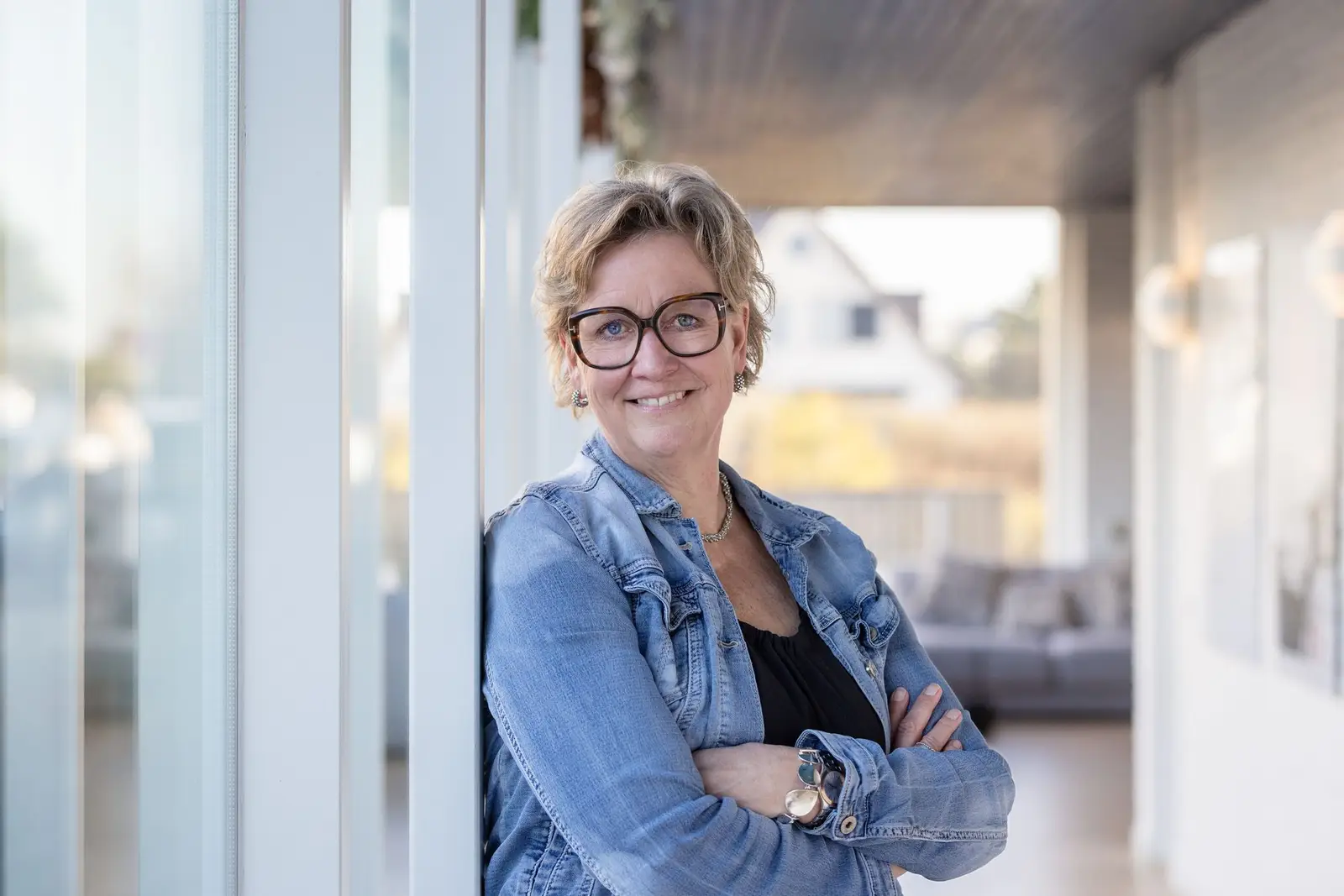 Een blonde vrouw met bril lachend kijkend in de camera leunend tegen een muur