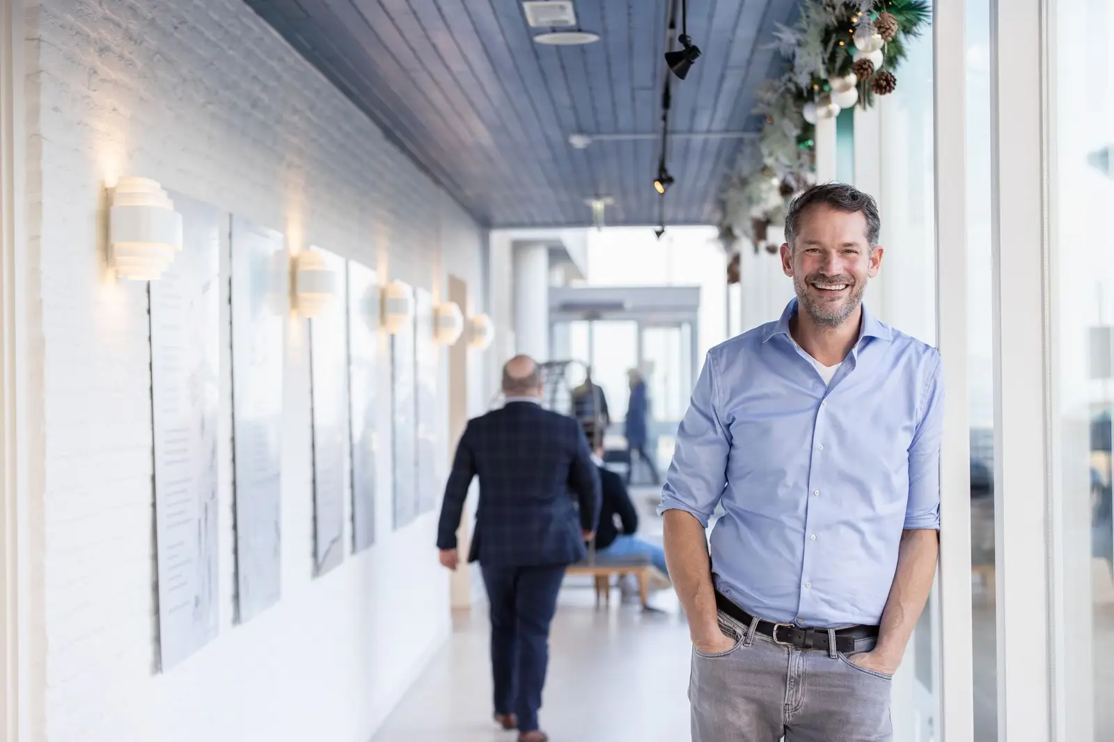Een vriendelijke man in blauwe blouse leunend tegen een muur in een gang